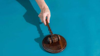 Mão segura martelo de juiz de madeira sobre base redonda de madeira, fundo azul.