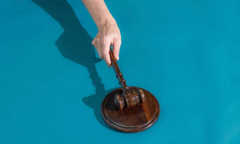 Mão segura martelo de juiz de madeira sobre base redonda de madeira, fundo azul.