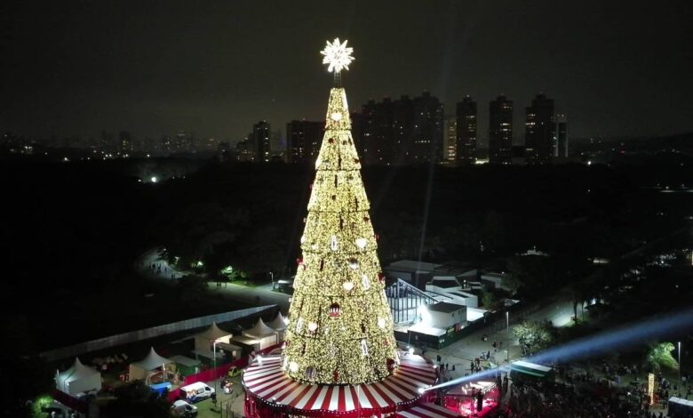 Árvore de Natal gigante decorada com luzes douradas e estrela no topo, situada em praça com tenda vermelha e branca na base. Multidão observa e circula ao redor durante a noite, com prédios ao fundo e iluminação pontual no ambiente.