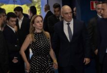 Homem de terno escuro e gravata azul caminha ao lado de mulher com vestido preto de bolinhas brancas, segurando as mãos. Ao redor, várias pessoas vestidas formalmente, algumas olhando para o casal. Ambiente interno com parede azul e sinal de saída de emergência.