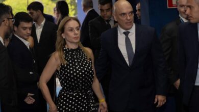 Homem de terno escuro e gravata azul caminha ao lado de mulher com vestido preto de bolinhas brancas, segurando as mãos. Ao redor, várias pessoas vestidas formalmente, algumas olhando para o casal. Ambiente interno com parede azul e sinal de saída de emergência.