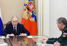 Dois homens sentados em lados opostos de uma mesa de reunião. À esquerda, homem de terno escuro com expressão séria, atrás dele bandeira russa com brasão. À direita, homem em uniforme militar verde com insígnias e bandeira da Rússia na manga, olhando para o interlocutor.