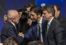 Dois homens de terno apertam as mãos em cumprimento durante evento formal. Um deles tem cabelo grisalho e barba branca, o outro tem cabelo escuro e barba curta. Ao fundo, fotógrafos registram o momento, e há outras pessoas em trajes formais.