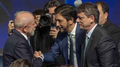 Dois homens de terno apertam as mãos em cumprimento durante evento formal. Um deles tem cabelo grisalho e barba branca, o outro tem cabelo escuro e barba curta. Ao fundo, fotógrafos registram o momento, e há outras pessoas em trajes formais.