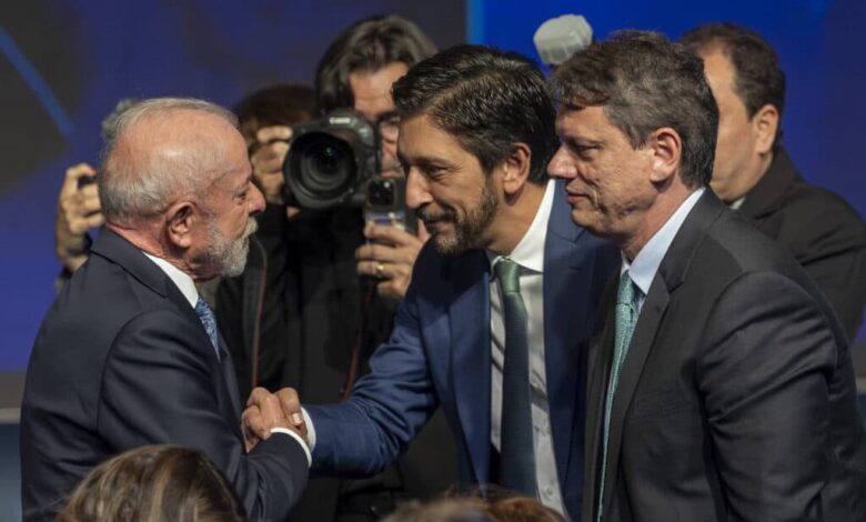 Dois homens de terno apertam as mãos em cumprimento durante evento formal. Um deles tem cabelo grisalho e barba branca, o outro tem cabelo escuro e barba curta. Ao fundo, fotógrafos registram o momento, e há outras pessoas em trajes formais.