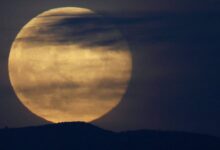 Lua cheia grande aparece parcialmente encoberta por nuvens finas e escuras, com silhueta de colina ao fundo em céu escuro.