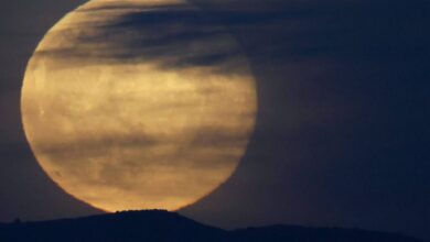 Lua cheia grande aparece parcialmente encoberta por nuvens finas e escuras, com silhueta de colina ao fundo em céu escuro.