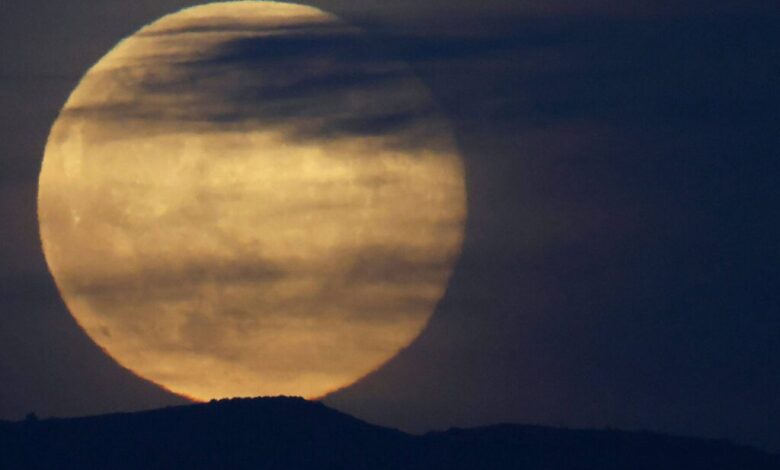 Lua cheia grande aparece parcialmente encoberta por nuvens finas e escuras, com silhueta de colina ao fundo em céu escuro.