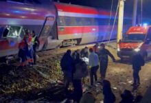 Trem com pintura vermelha, branca e azul parado nos trilhos à noite. Pessoas e equipes de resgate estão próximas ao trem e a uma ambulância com luzes ligadas.