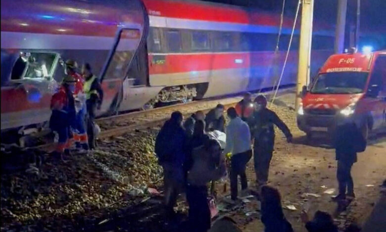 Trem com pintura vermelha, branca e azul parado nos trilhos à noite. Pessoas e equipes de resgate estão próximas ao trem e a uma ambulância com luzes ligadas.