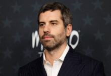 Homem de cabelos curtos e barba aparada veste terno escuro listrado e camisa branca, posando diante de fundo escuro com estrelas e texto desfocado.