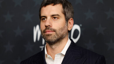 Homem de cabelos curtos e barba aparada veste terno escuro listrado e camisa branca, posando diante de fundo escuro com estrelas e texto desfocado.