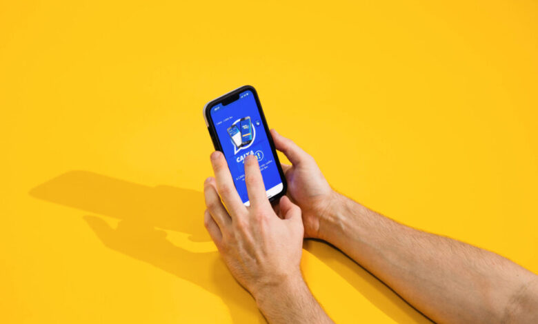 A imagem mostra duas mãos segurando um smartphone com a tela voltada para cima. O smartphone exibe um aplicativo com um fundo azul e um ícone central. O fundo da imagem é amarelo, criando um contraste vibrante.