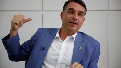 Homem de meia-idade veste terno azul e camisa branca, sentado em ambiente interno com parede branca ao fundo. Ele gesticula com a mão esquerda apontando para trás e segura um copo com a mão direita.