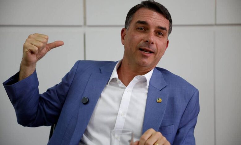 Homem de meia-idade veste terno azul e camisa branca, sentado em ambiente interno com parede branca ao fundo. Ele gesticula com a mão esquerda apontando para trás e segura um copo com a mão direita.