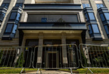 Entrada de prédio com colunas e vidro, protegida por barreiras metálicas alinhadas na calçada. Fachada moderna com janelas espelhadas e o nome