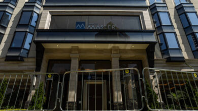 Entrada de prédio com colunas e vidro, protegida por barreiras metálicas alinhadas na calçada. Fachada moderna com janelas espelhadas e o nome