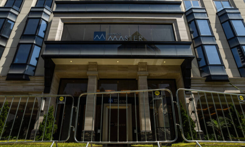 Entrada de prédio com colunas e vidro, protegida por barreiras metálicas alinhadas na calçada. Fachada moderna com janelas espelhadas e o nome