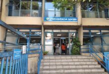 A imagem mostra a entrada de uma agência da Previdência Social. A fachada é de um prédio de dois andares, com janelas grandes e uma escada de acesso. Acima da entrada, há uma placa azul com o texto