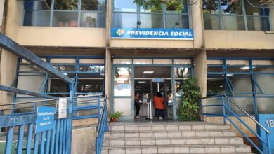 A imagem mostra a entrada de uma agência da Previdência Social. A fachada é de um prédio de dois andares, com janelas grandes e uma escada de acesso. Acima da entrada, há uma placa azul com o texto