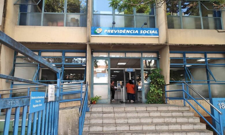 A imagem mostra a entrada de uma agência da Previdência Social. A fachada é de um prédio de dois andares, com janelas grandes e uma escada de acesso. Acima da entrada, há uma placa azul com o texto