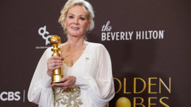 Mulher loira em vestido branco com detalhes dourados segura estatueta do Globo de Ouro em frente a fundo com logos do Beverly Hilton e Golden Globes.