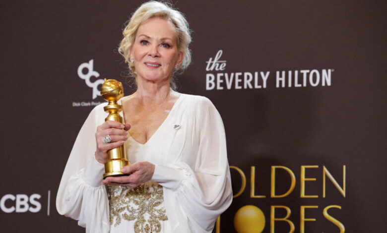 Mulher loira em vestido branco com detalhes dourados segura estatueta do Globo de Ouro em frente a fundo com logos do Beverly Hilton e Golden Globes.