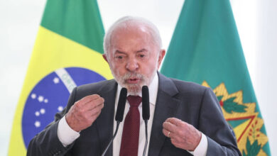 Homem de terno e gravata fala em microfones com bandeiras do Brasil e outra institucional ao fundo.