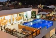 Piscina retangular iluminada em azul ocupa o centro da cobertura de prédio residencial. Ao redor, há guarda-corpos de vidro e áreas com mesas e cadeiras. Ambiente interno adjacente com iluminação amarela e móveis de madeira é visível à esquerda. Ao fundo, casas e edifícios baixos sob céu noturno.