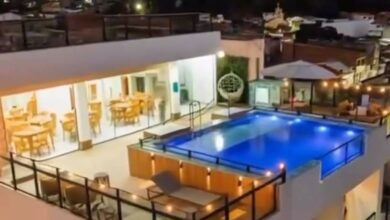 Piscina retangular iluminada em azul ocupa o centro da cobertura de prédio residencial. Ao redor, há guarda-corpos de vidro e áreas com mesas e cadeiras. Ambiente interno adjacente com iluminação amarela e móveis de madeira é visível à esquerda. Ao fundo, casas e edifícios baixos sob céu noturno.