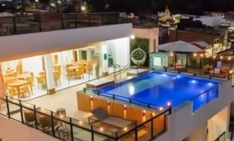 Piscina retangular iluminada em azul ocupa o centro da cobertura de prédio residencial. Ao redor, há guarda-corpos de vidro e áreas com mesas e cadeiras. Ambiente interno adjacente com iluminação amarela e móveis de madeira é visível à esquerda. Ao fundo, casas e edifícios baixos sob céu noturno.
