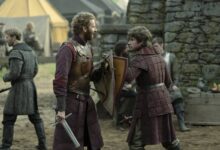 Dois homens vestidos com armaduras e roupas medievais conversam em um acampamento ao ar livre. Um deles segura uma espada e um escudo, enquanto o outro veste uma armadura acolchoada. Ao fundo, há tendas e outras pessoas vestidas de forma semelhante, além de uma parede de pedra.