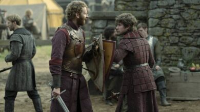 Dois homens vestidos com armaduras e roupas medievais conversam em um acampamento ao ar livre. Um deles segura uma espada e um escudo, enquanto o outro veste uma armadura acolchoada. Ao fundo, há tendas e outras pessoas vestidas de forma semelhante, além de uma parede de pedra.