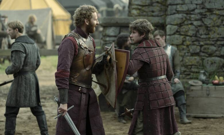 Dois homens vestidos com armaduras e roupas medievais conversam em um acampamento ao ar livre. Um deles segura uma espada e um escudo, enquanto o outro veste uma armadura acolchoada. Ao fundo, há tendas e outras pessoas vestidas de forma semelhante, além de uma parede de pedra.