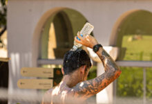 Homem sem camisa com tatuagens no braço e relógio no pulso despeja água de uma garrafa plástica na cabeça. Ao fundo, estrutura com arcos e placas de sinalização indicam ambiente externo.