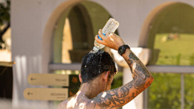 Homem sem camisa com tatuagens no braço e relógio no pulso despeja água de uma garrafa plástica na cabeça. Ao fundo, estrutura com arcos e placas de sinalização indicam ambiente externo.