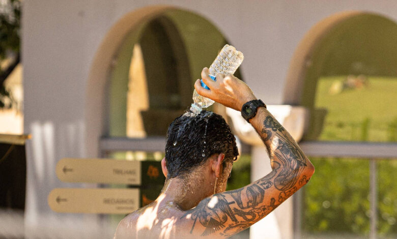 Homem sem camisa com tatuagens no braço e relógio no pulso despeja água de uma garrafa plástica na cabeça. Ao fundo, estrutura com arcos e placas de sinalização indicam ambiente externo.