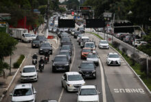 Vários veículos, incluindo carros e motos, formam congestionamento em avenida de pista dupla com faixa exclusiva para ônibus à direita. Árvores e construções cercam a via em dia nublado.