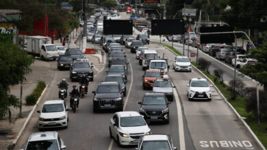 Vários veículos, incluindo carros e motos, formam congestionamento em avenida de pista dupla com faixa exclusiva para ônibus à direita. Árvores e construções cercam a via em dia nublado.