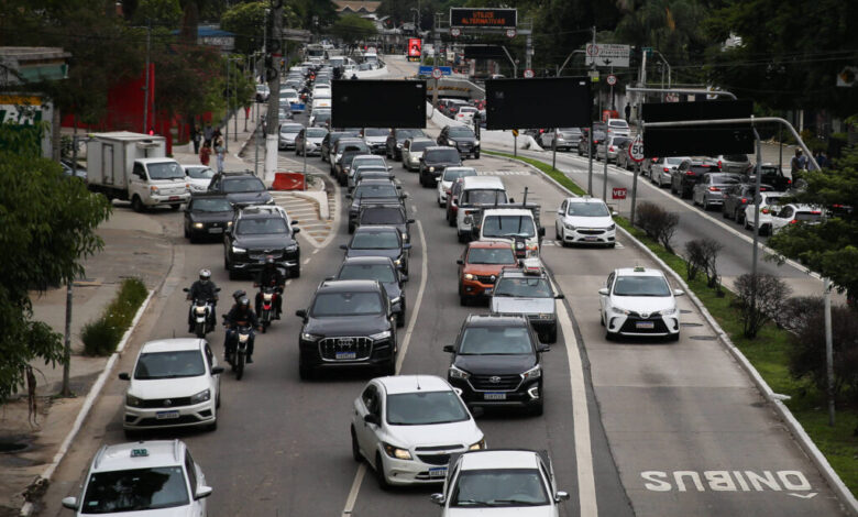 Vários veículos, incluindo carros e motos, formam congestionamento em avenida de pista dupla com faixa exclusiva para ônibus à direita. Árvores e construções cercam a via em dia nublado.