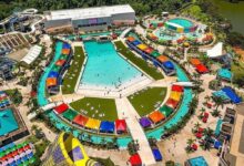 Vista aérea de parque aquático com piscina central grande em formato hexagonal, cercada por tendas coloridas e áreas gramadas com espreguiçadeiras. Ao redor, toboáguas, piscina circular menor, espreguiçadeiras com guarda-sóis coloridos e caminhos pavimentados. Vegetação e lago ao fundo.