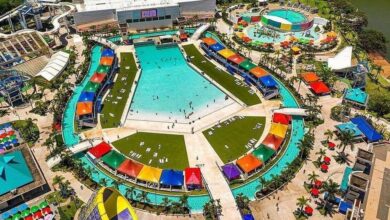 Vista aérea de parque aquático com piscina central grande em formato hexagonal, cercada por tendas coloridas e áreas gramadas com espreguiçadeiras. Ao redor, toboáguas, piscina circular menor, espreguiçadeiras com guarda-sóis coloridos e caminhos pavimentados. Vegetação e lago ao fundo.