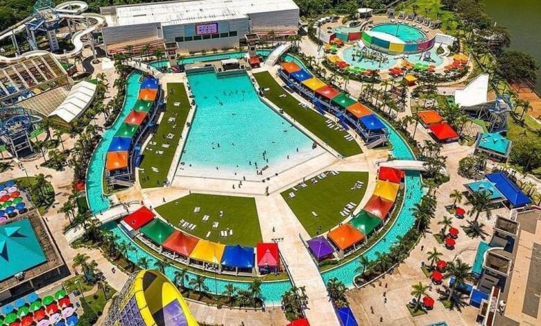 Vista aérea de parque aquático com piscina central grande em formato hexagonal, cercada por tendas coloridas e áreas gramadas com espreguiçadeiras. Ao redor, toboáguas, piscina circular menor, espreguiçadeiras com guarda-sóis coloridos e caminhos pavimentados. Vegetação e lago ao fundo.